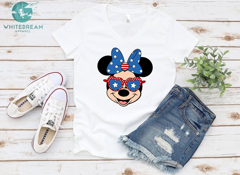 May include: White V-neck t-shirt with a red, white, and blue Minnie Mouse graphic wearing heart-shaped sunglasses. The graphic features a red bow with white stars and a red, white, and blue bow. The shirt is laying on a white wooden surface with a pair of white Converse shoes and a pair of blue denim shorts.