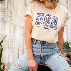 May include: Cream-colored t-shirt with the word "USA" in a floral design and the text "EST. 1776" in red. The shirt is paired with light blue denim jeans. The overall style is casual and patriotic.