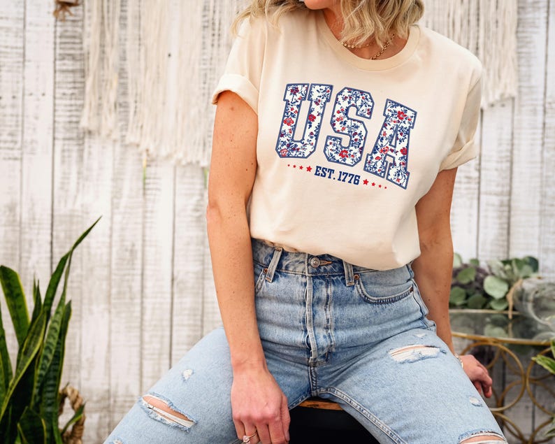 May include: Cream-colored t-shirt with the word "USA" in a floral design and the text "EST. 1776" in red. The shirt is paired with light blue denim jeans. The overall style is casual and patriotic.