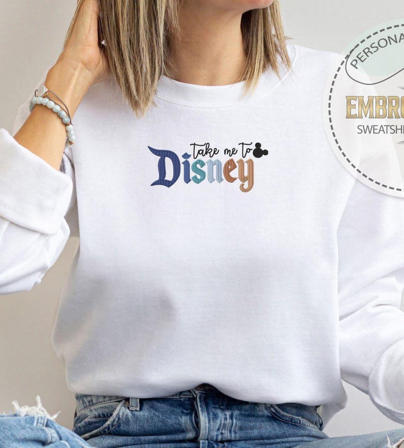 Puede incluir: Sudadera blanca con la frase bordada "Take me to Disney" en azul, turquesa y marr&oacute;n. El dise&ntilde;o incluye una cabeza de Mickey Mouse. La sudadera es de estilo casual.