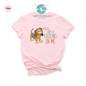 Op de afbeelding: Roze T-shirt met een cartoon afbeelding van een hond uit de Toy Story-filmfranchise. De tekst "You've Got a Friend in Me" staat op het shirt gedrukt.