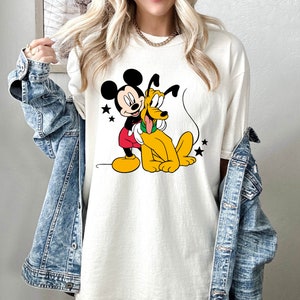 May include: White t-shirt with a graphic print of Mickey Mouse and Pluto. Mickey Mouse is wearing red shorts and yellow shoes. Pluto is wearing a green collar. The graphic is surrounded by stars.