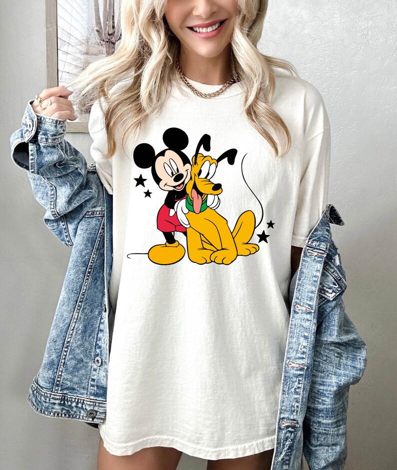 May include: White t-shirt with a graphic print of Mickey Mouse and Pluto. Mickey Mouse is wearing red shorts and yellow shoes. Pluto is wearing a green collar. The graphic is surrounded by stars.