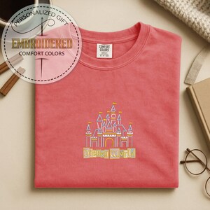 May include: Coral-colored Comfort Colors t-shirt with an embroidered Disney World castle design. The castle features yellow, purple, and blue accents, with the words "Disney World" embroidered below. The shirt also has a tag that says "Comfort Colors".
