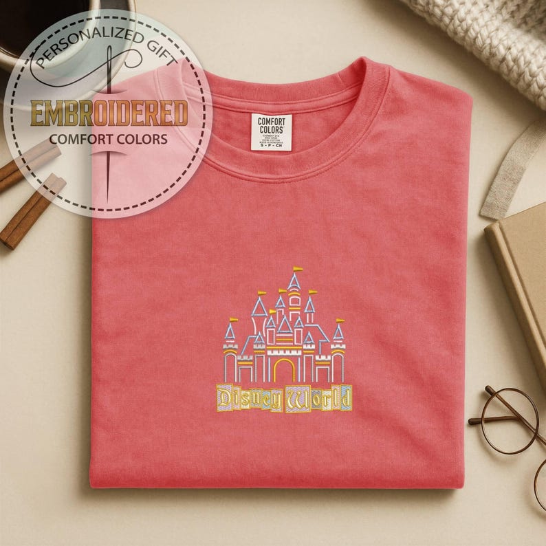 May include: Coral-colored Comfort Colors t-shirt with an embroidered Disney World castle design. The castle features yellow, purple, and blue accents, with the words "Disney World" embroidered below. The shirt also has a tag that says "Comfort Colors".