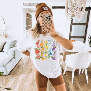 May include: White t-shirt with a rainbow floral print. The shirt has a crew neck and short sleeves. The model is wearing brown shorts and a brown beanie with the text "Good Vibes" on it.