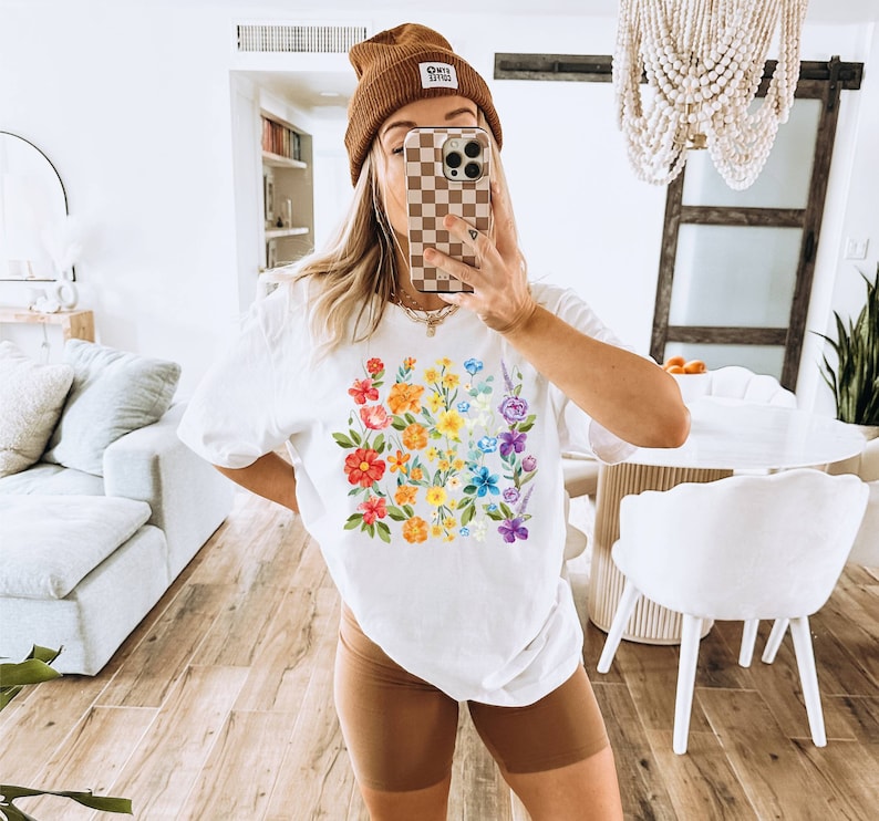 May include: White t-shirt with a rainbow floral print. The shirt has a crew neck and short sleeves. The model is wearing brown shorts and a brown beanie with the text "Good Vibes" on it.