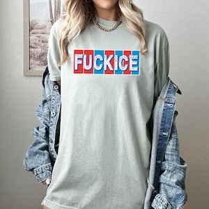 以下が含まれることがあります： 赤、白、青のブロック文字で「FUCKICE」と書かれたセージグリーンのTシャツ。 ゆったりとしたフィット感のシャツで、デニムジャケットと合わせて着用されています。