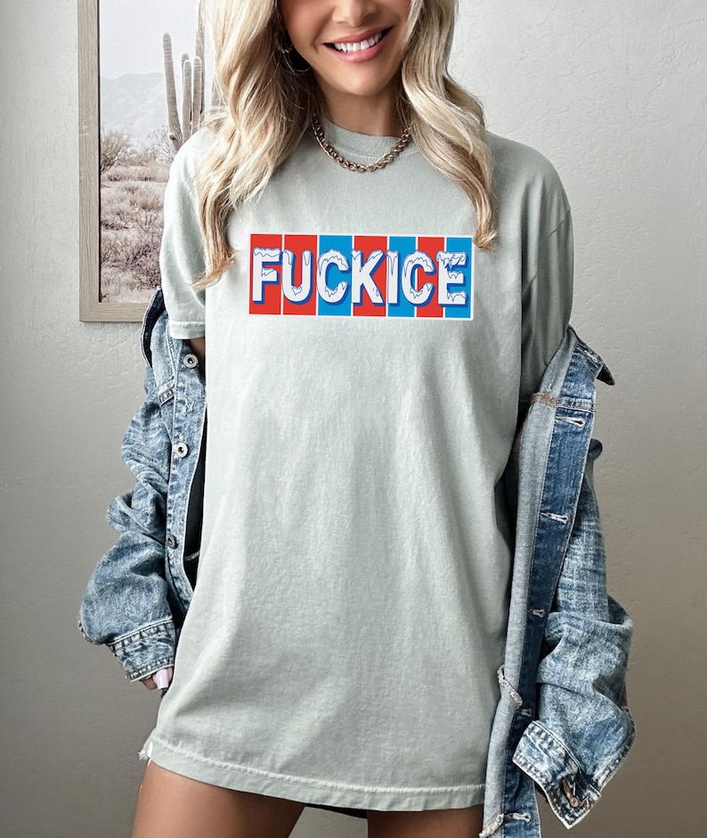 以下が含まれることがあります： 赤、白、青のブロック文字で「FUCKICE」と書かれたセージグリーンのTシャツ。 ゆったりとしたフィット感のシャツで、デニムジャケットと合わせて着用されています。