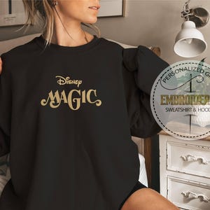 Peut inclure: Sweat-shirt noir avec l'inscription dor&eacute;e "Disney Magic" brod&eacute;e. Ce haut &agrave; manches longues et col rond pr&eacute;sente un motif circulaire sur le c&ocirc;t&eacute; droit avec les mots "Personalized Gift" et "Embroidered Sweatshirt & Hoodie".