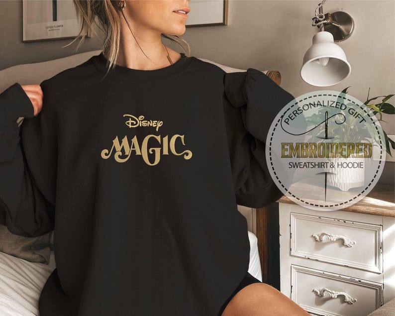 Peut inclure: Sweat-shirt noir avec l'inscription dor&eacute;e "Disney Magic" brod&eacute;e. Ce haut &agrave; manches longues et col rond pr&eacute;sente un motif circulaire sur le c&ocirc;t&eacute; droit avec les mots "Personalized Gift" et "Embroidered Sweatshirt & Hoodie".