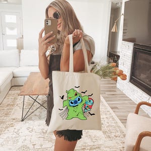 Pode incluir: Uma sacola creme com um personagem de desenho animado verde com um rosto azul, segurando um personagem menor. Morcegos pretos e teias de aranha decoram a sacola. A sacola tem duas al&ccedil;as longas.