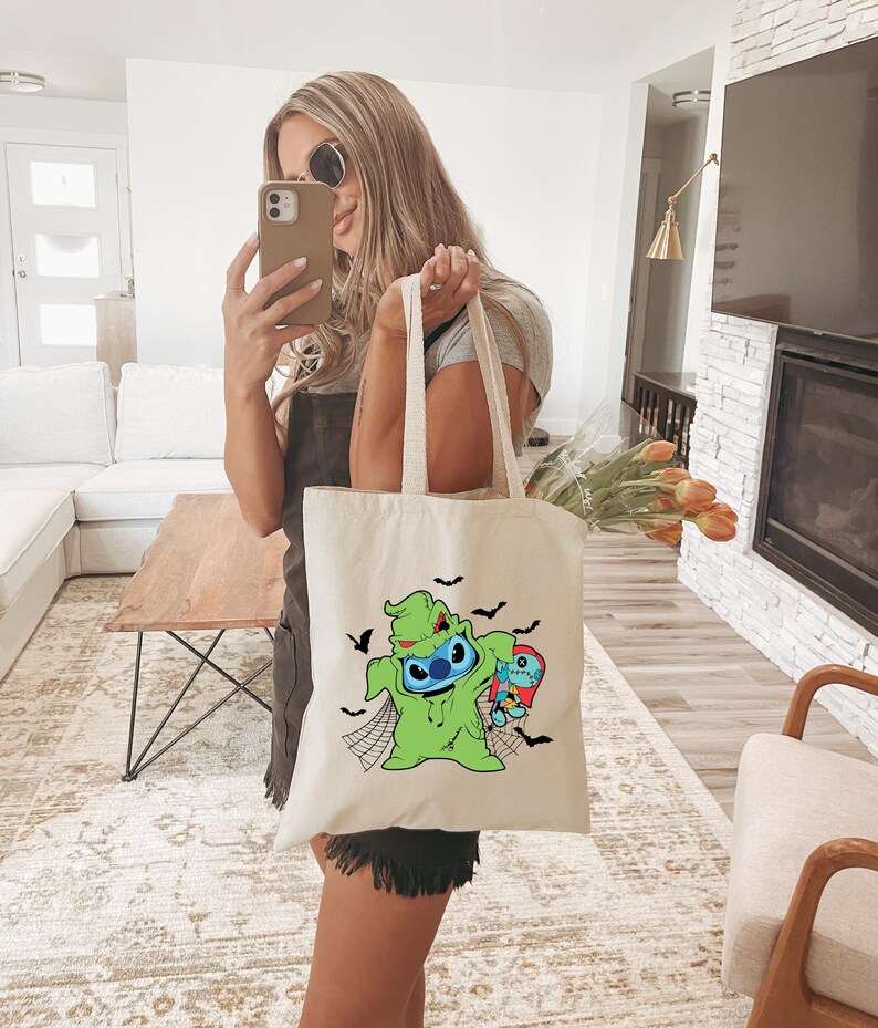 Pode incluir: Uma sacola creme com um personagem de desenho animado verde com um rosto azul, segurando um personagem menor. Morcegos pretos e teias de aranha decoram a sacola. A sacola tem duas al&ccedil;as longas.