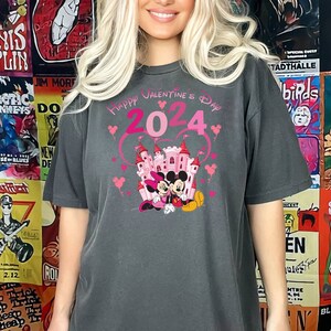 May include: A dark gray t-shirt with a pink graphic of Mickey Mouse and Minnie Mouse in front of a pink castle. The graphic also includes the text "Happy Valentine's Day 2024" in pink.