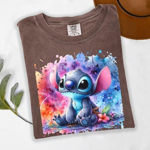 Pu&ograve; includere: Una maglietta Comfort Colors marrone con una grafica ad acquerello di Stitch di Lilo & Stitch. Il design mostra Stitch con grandi occhi e orecchie, su uno sfondo vibrante di blu, rosa e arancione.