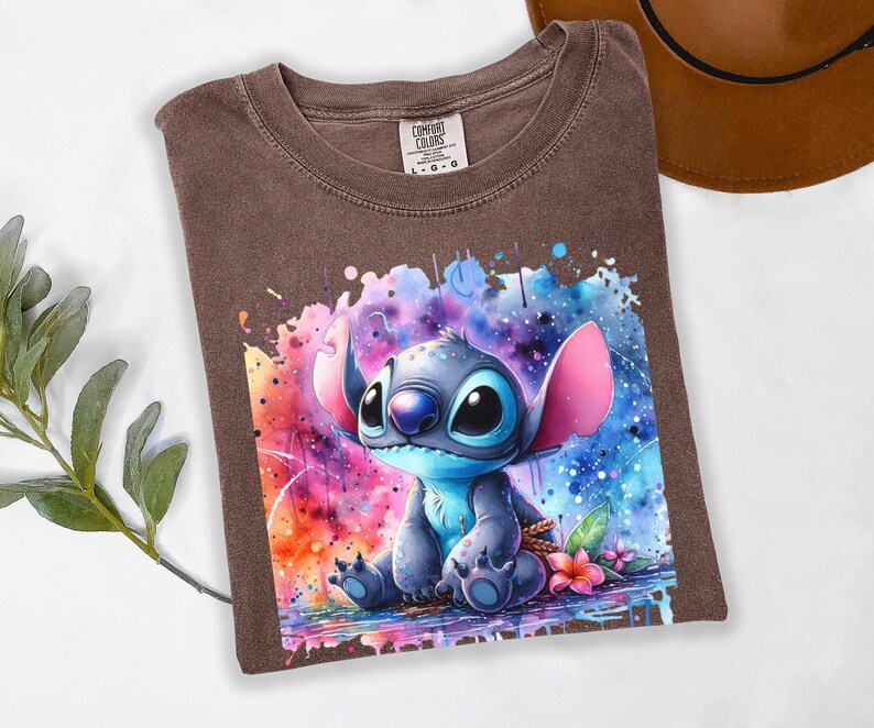 Pu&ograve; includere: Una maglietta Comfort Colors marrone con una grafica ad acquerello di Stitch di Lilo & Stitch. Il design mostra Stitch con grandi occhi e orecchie, su uno sfondo vibrante di blu, rosa e arancione.