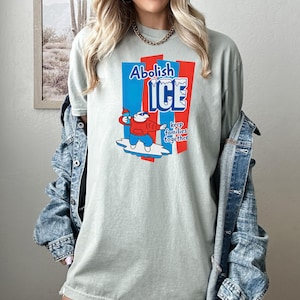 May include: A light green t-shirt with the words "Abolish ICE" in blue and red, along with a cartoon character holding a popsicle. The text "keep families together" is also included. The shirt is a casual style.