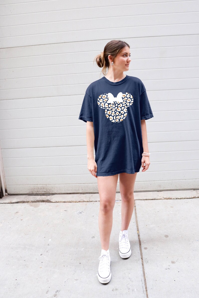 Peut inclure: Un t-shirt bleu marine avec un contour blanc de la t&ecirc;te de Mickey Mouse, rempli d'un motif l&eacute;opard en or et blanc. Le n&oelig;ud sur la t&ecirc;te de Mickey est blanc.