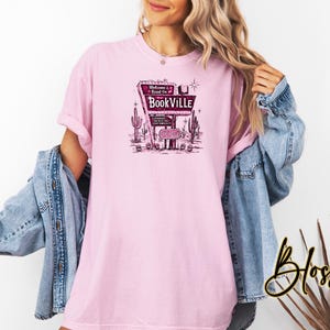 May include: Pink t-shirt with a graphic of a vintage motel sign that says "Welcome to Bookville". The sign is surrounded by cacti and stars.