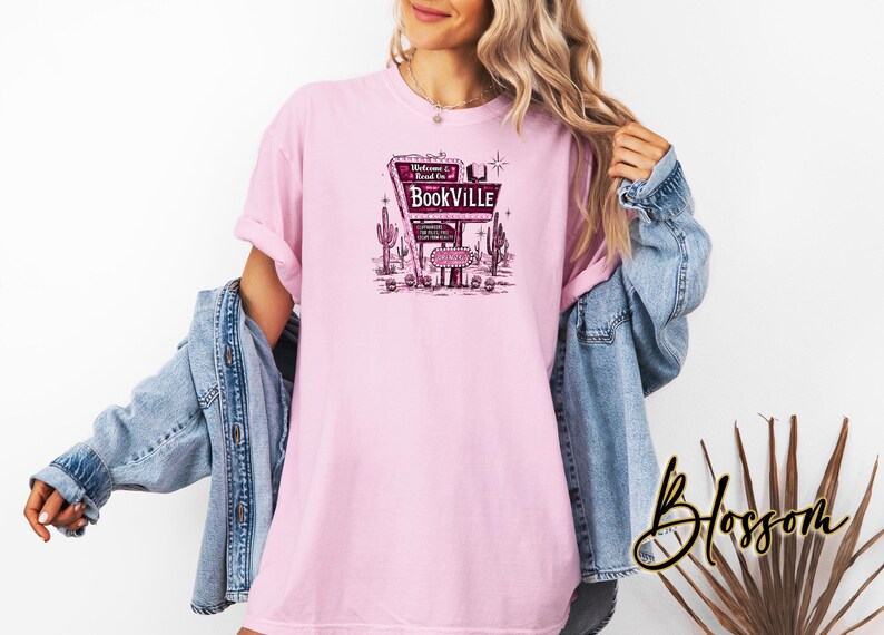May include: Pink t-shirt with a graphic of a vintage motel sign that says "Welcome to Bookville". The sign is surrounded by cacti and stars.