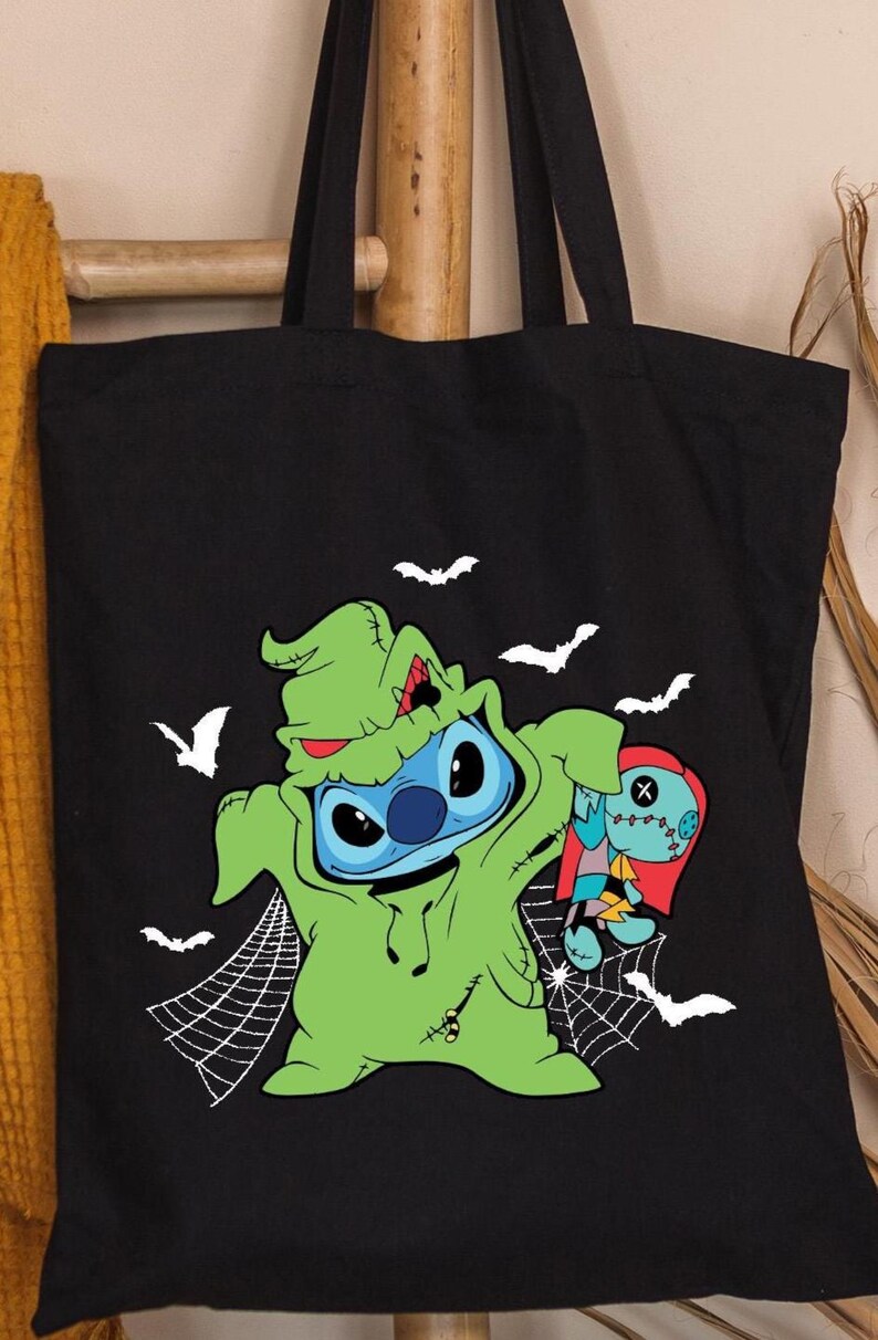 Pode incluir: Bolsa tote preta com um design de Halloween. Apresenta um personagem azul com uma fantasia verde, segurando uma boneca, com morcegos brancos e teias de aranha. A bolsa &eacute; feita de tecido dur&aacute;vel.