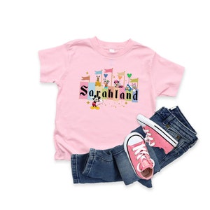 以下が含まれることがあります： ピンクのTシャツに、ミッキーマウスと他のディズニーキャラクターが城の前で「SarahLand」という文字が書かれたカラフルなグラフィックがプリントされています。