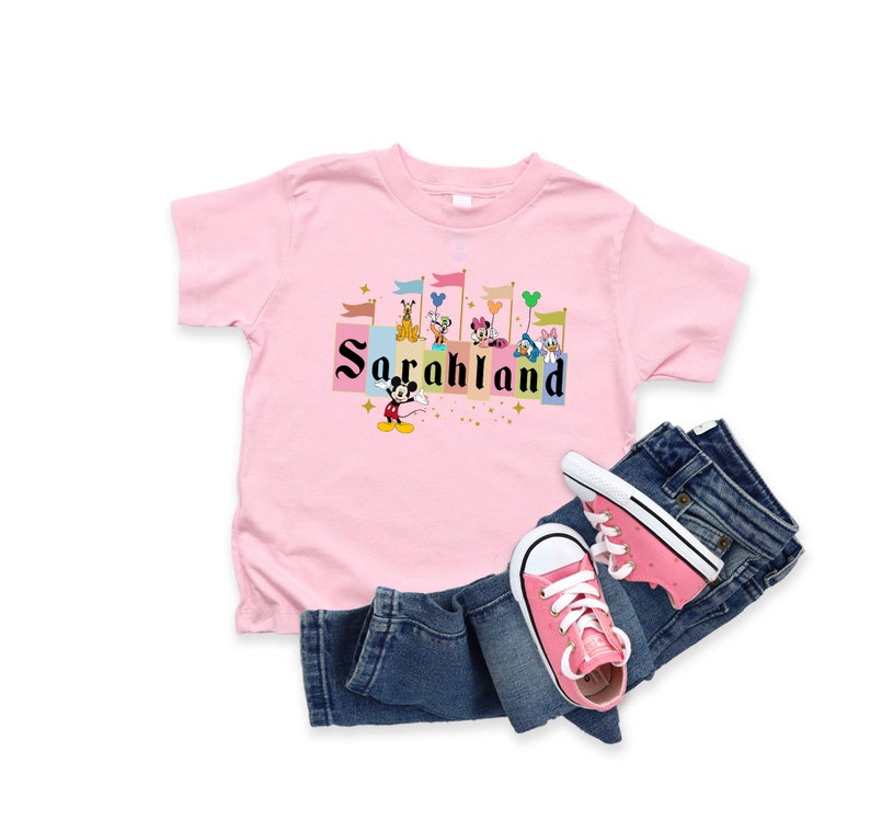 以下が含まれることがあります： ピンクのTシャツに、ミッキーマウスと他のディズニーキャラクターが城の前で「SarahLand」という文字が書かれたカラフルなグラフィックがプリントされています。