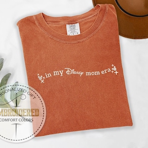 May include: A rust-colored t-shirt with the text "in my Disney mom era" embroidered in white. The text is accompanied by two small white Mickey Mouse heads.