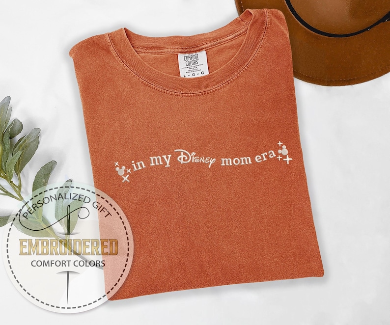 May include: A rust-colored t-shirt with the text "in my Disney mom era" embroidered in white. The text is accompanied by two small white Mickey Mouse heads.