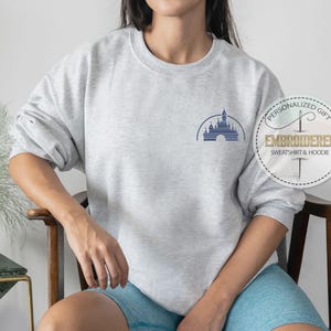 May include: A gray sweatshirt with a blue embroidered image of the Disney castle. The sweatshirt is being worn by a person. The text "Personalized Gift" and "Embroidered Sweatshirt & Hoodie" is on a white tag.
