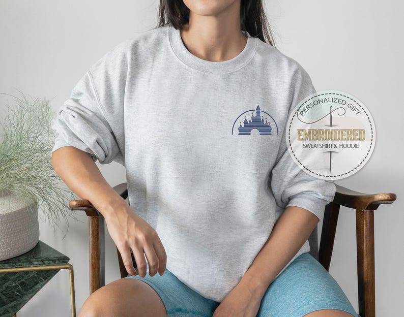 May include: A gray sweatshirt with a blue embroidered image of the Disney castle. The sweatshirt is being worn by a person. The text "Personalized Gift" and "Embroidered Sweatshirt & Hoodie" is on a white tag.