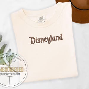 以下が含まれることがあります： クリーム色のTシャツに、茶色の筆記体で「Disneyland」と刺繍されています。このシャツはComfort Colorsブランドで、パーソナライズされたギフトタグも含まれています。