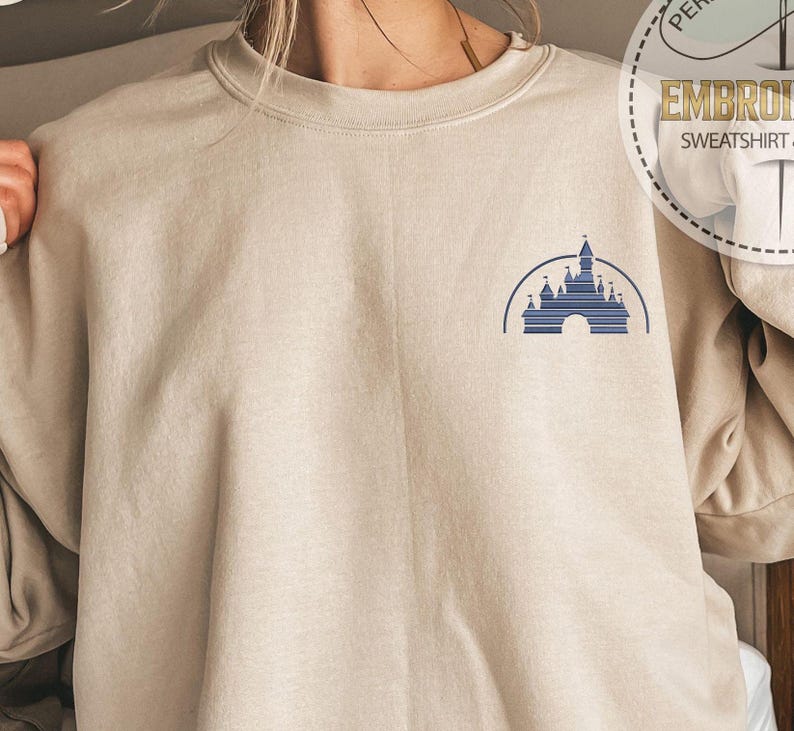 May include: A beige sweatshirt with a blue embroidered image of a castle on the left chest. The embroidery is of a castle with a pointed roof and a large archway. The castle is surrounded by a blue arch.