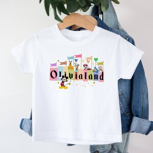 以下が含まれることがあります： 白いTシャツに、ミッキーマウスとその仲間たちが城の前で「Olivia Land」と書かれたカラフルなグラフィックがプリントされています。