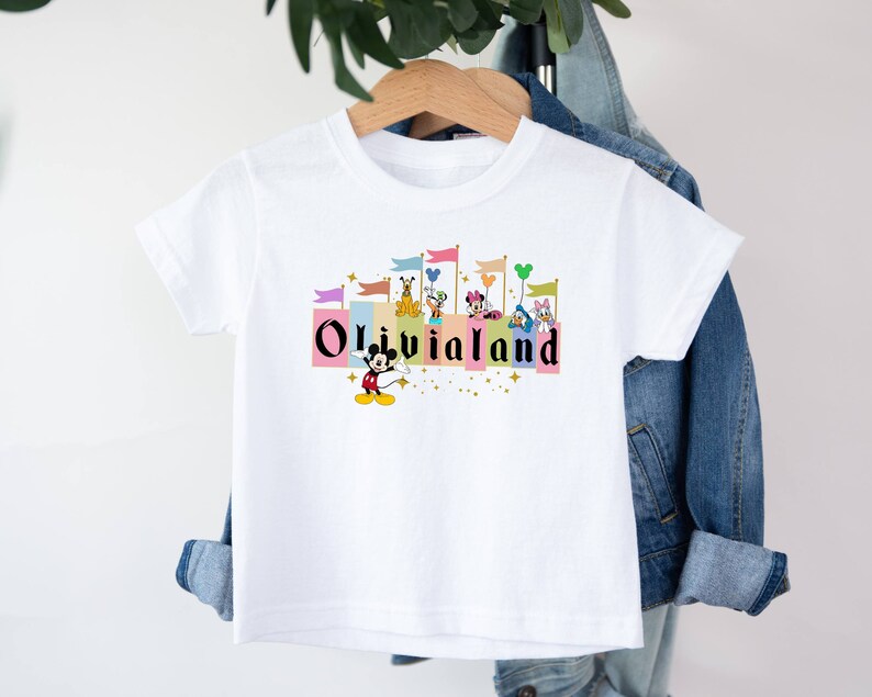 以下が含まれることがあります： 白いTシャツに、ミッキーマウスとその仲間たちが城の前で「Olivia Land」と書かれたカラフルなグラフィックがプリントされています。