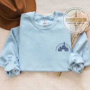 May include: Light blue sweatshirt with a small embroidered image of Cinderella's castle on the left chest. The sweatshirt is folded and laid flat on a light brown surface.