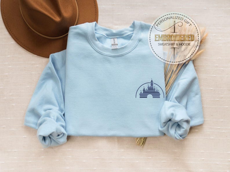 May include: Light blue sweatshirt with a small embroidered image of Cinderella's castle on the left chest. The sweatshirt is folded and laid flat on a light brown surface.