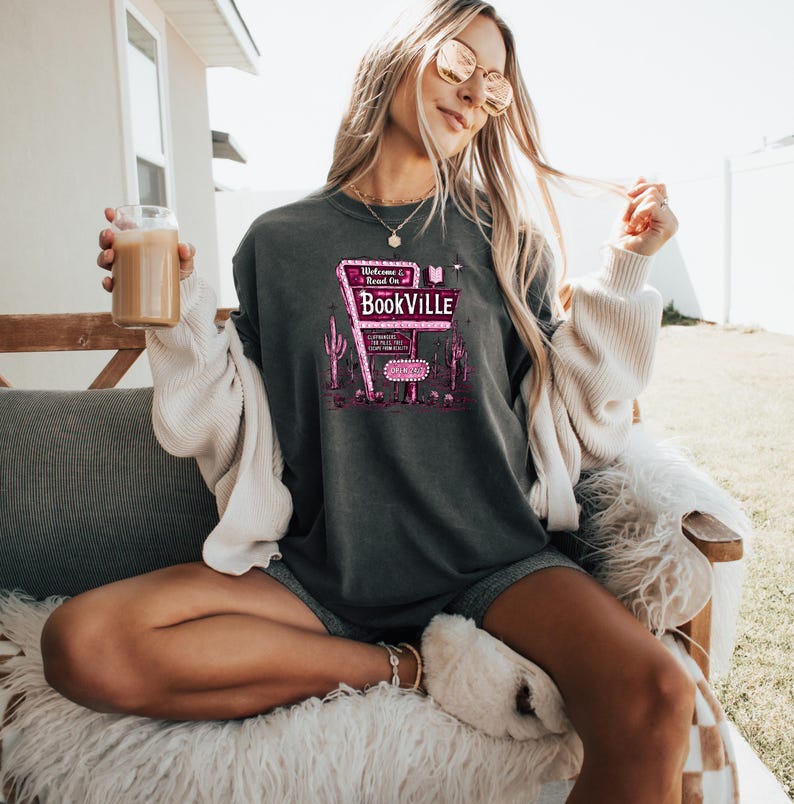 May include: A dark gray t-shirt with a pink graphic of a neon sign that reads "Welcome to Bookville" with a book icon. The sign is on a desert background with cacti.