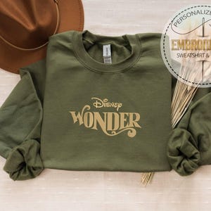 Peut inclure: Un sweat-shirt vert olive avec le texte "Disney Wonder" en lettres dor&eacute;es. Un chapeau marron est visible dans le coin sup&eacute;rieur gauche. Le sweat-shirt fait partie d'une collection de cadeaux brod&eacute;s personnalis&eacute;s.