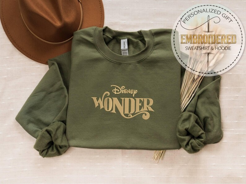 Peut inclure: Un sweat-shirt vert olive avec le texte "Disney Wonder" en lettres dor&eacute;es. Un chapeau marron est visible dans le coin sup&eacute;rieur gauche. Le sweat-shirt fait partie d'une collection de cadeaux brod&eacute;s personnalis&eacute;s.