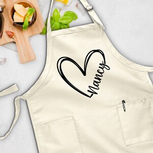 May include: A white apron with a black heart and the name "Nancy" written inside. The apron has a pocket on the front.