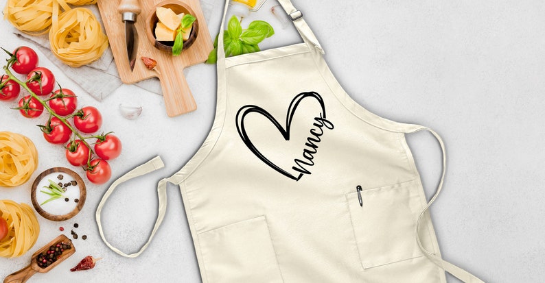 May include: A white apron with a black heart and the name "Nancy" written inside. The apron has a pocket on the front.