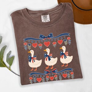 May include: Brown Comfort Colors t-shirt featuring a patriotic design with three cartoon ducks wearing bow ties with red, white, and blue accents. The shirt also has red hearts and blue bows.