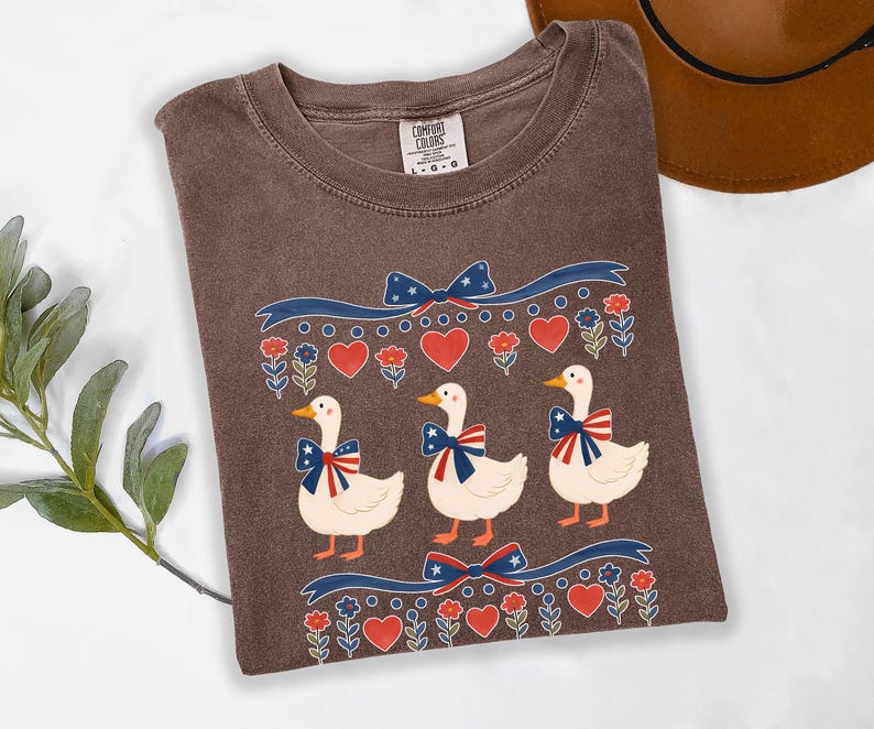 May include: Brown Comfort Colors t-shirt featuring a patriotic design with three cartoon ducks wearing bow ties with red, white, and blue accents. The shirt also has red hearts and blue bows.