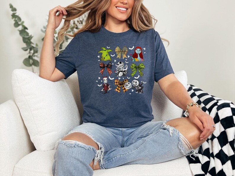 May include: Navy blue t-shirt featuring a colorful graphic of characters and bows from The Nightmare Before Christmas. The design includes images of Sally, Jack Skellington, and Oogie Boogie. The shirt is a crew neck and has short sleeves.