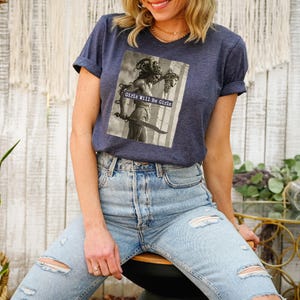 May include: Navy blue t-shirt featuring a sepia-toned graphic of a statue holding a sword, with the text "Girls Will Be Girls." The shirt has rolled-up sleeves and is paired with light-wash denim jeans.