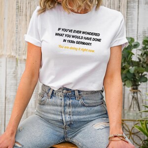 May include: White t-shirt with black text that reads "IF YOU'VE EVER WONDERED WHAT YOU WOULD HAVE DONE IN 1930s GERMANY: You are doing it right now."