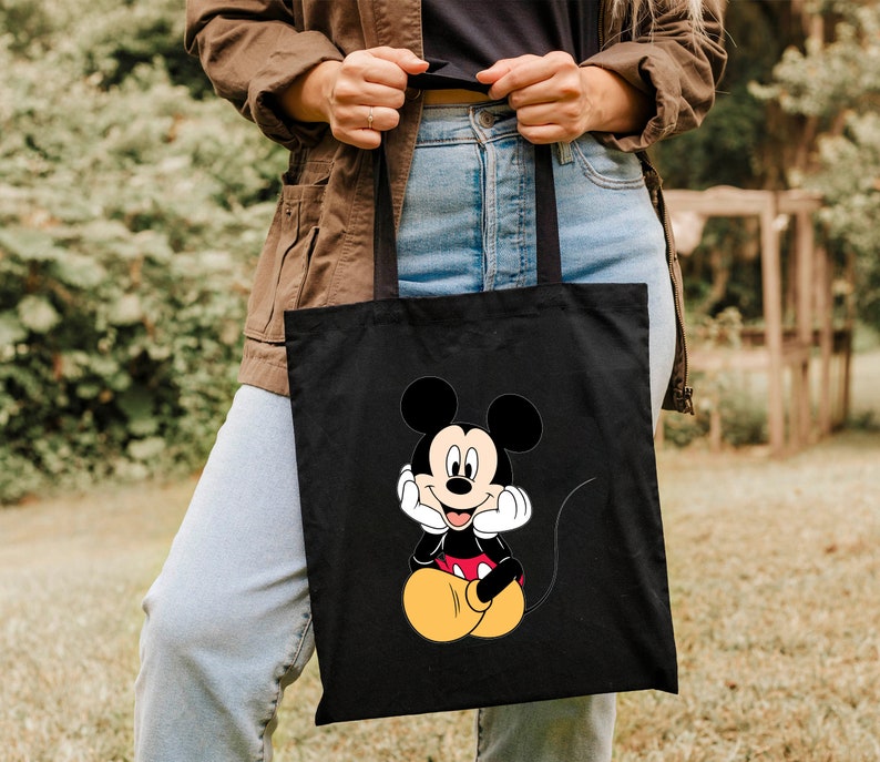 K&ouml;nnte beinhalten: Schwarze Tragetasche mit einem Cartoon-Bild von Mickey Mouse in einem roten und gelben Outfit. Mickey Mouse sitzt mit den H&auml;nden am Kinn und hat ein breites L&auml;cheln.