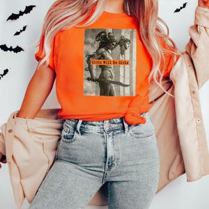 May include: Orange t-shirt featuring a sepia-toned graphic of a statue of Medusa holding a sword and the severed head of a woman. The text "Girls Will Be Girls" is printed on the graphic. The model is wearing light wash jeans and a tan jacket.