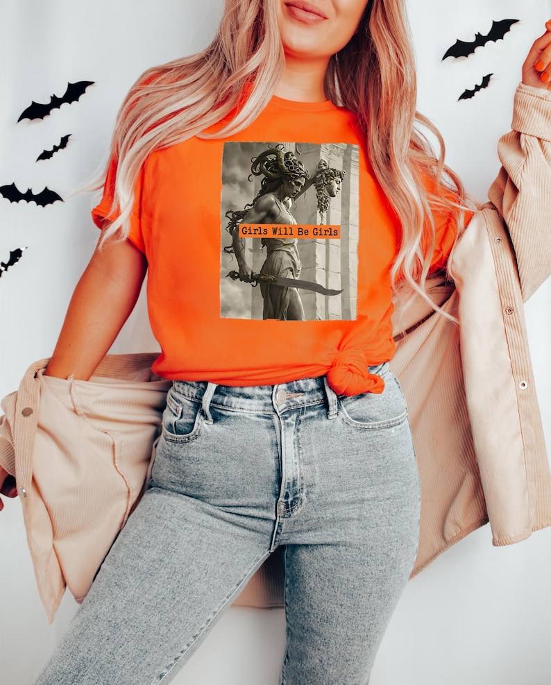 May include: Orange t-shirt featuring a sepia-toned graphic of a statue of Medusa holding a sword and the severed head of a woman. The text "Girls Will Be Girls" is printed on the graphic. The model is wearing light wash jeans and a tan jacket.
